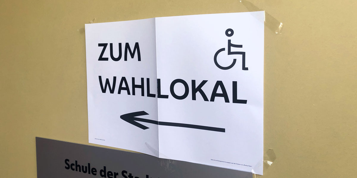 Zettel an der Wand "zum Wahllokal" mit Pfeil und Rollstuhl-Symbol